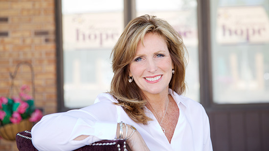 Susan Bertrand sits outside smiling and facing the camera. She is wearing a white blouse and silver jewelry. Behind her are 3 large window panes, each displaying the Maureen’s Hope Foundation logo, next to a brick wall and a flowering potted plant. 